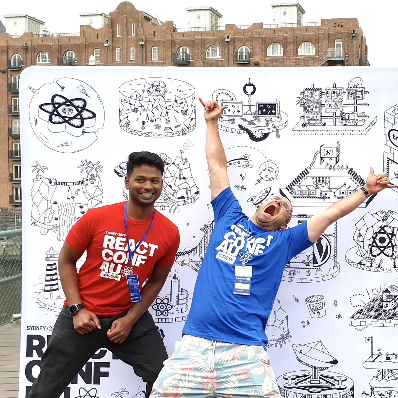 Dinesh Pandiyan & Dominik Wilkowski having fun at ReactConf AU in front of a ReactConf AU banner poster