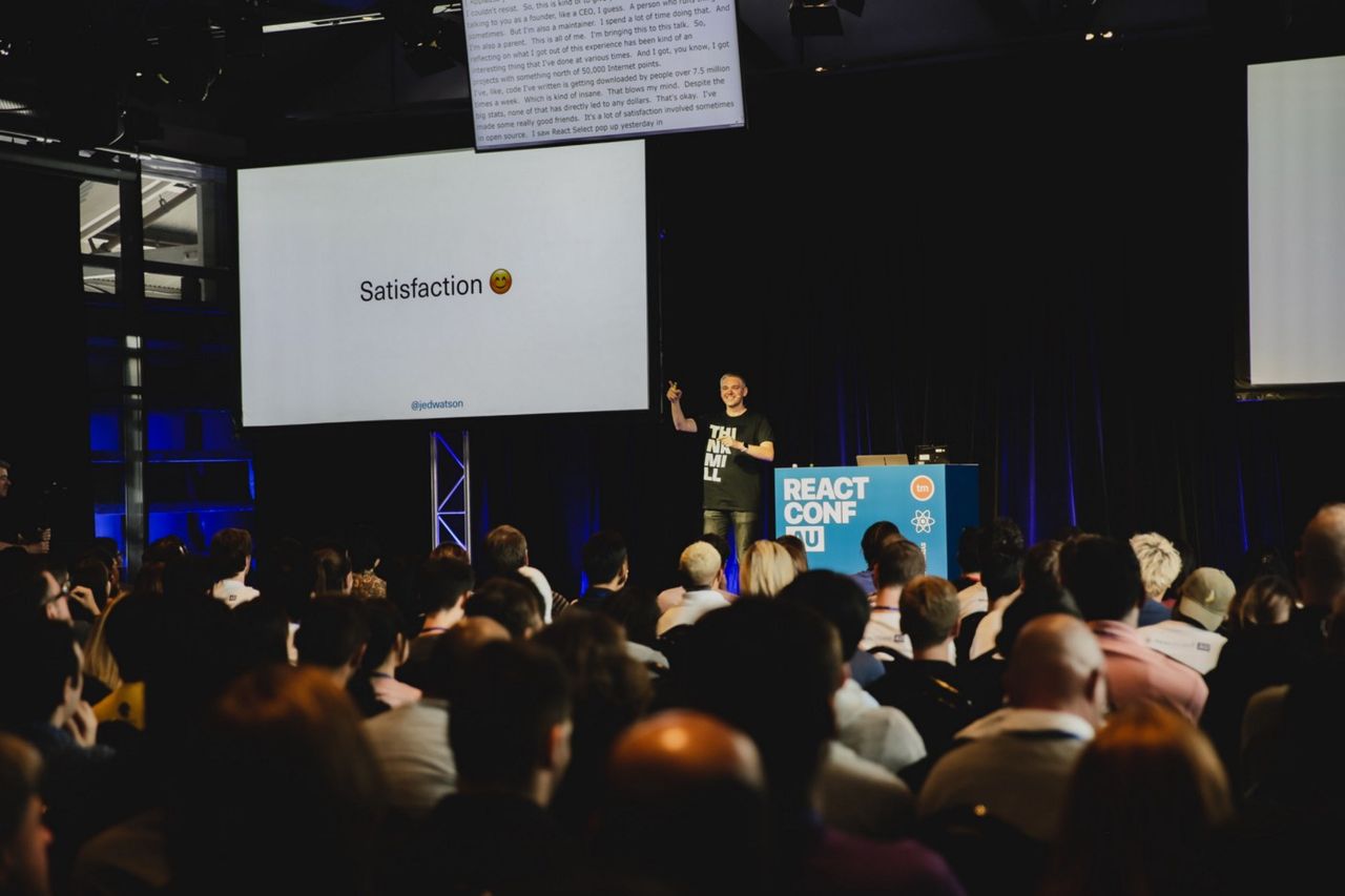 Jed Watson presenting on stage at ReactConf AU with a screen next to him displaying 'Satisfaction ☺️'